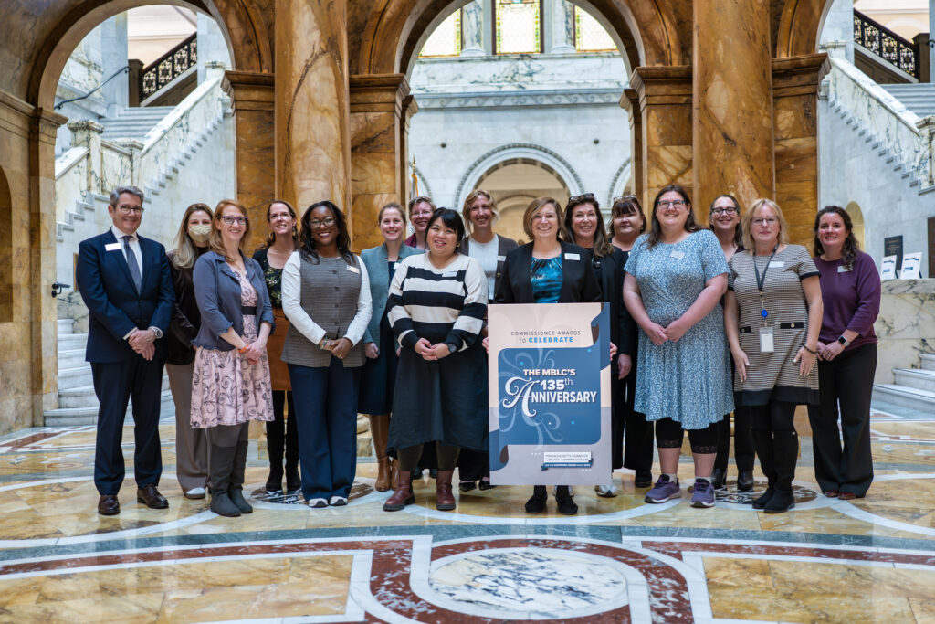 Group photo of the MBLC Staff at the Commissioner Awards on November 6, 2025, at the Massachusetts State House, posing behind a sign that reads “The MBLC’s 135th Anniversary".
