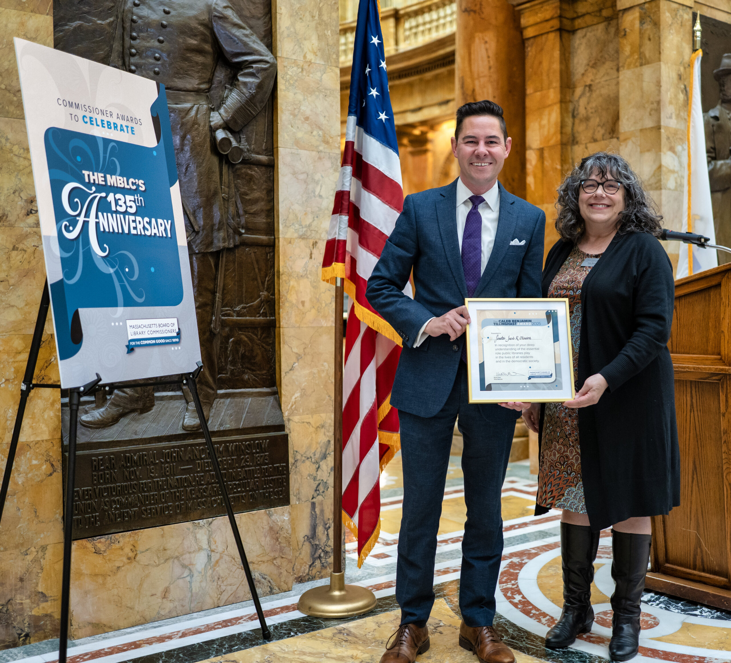 A man and woman stand indoors at the 2025 Commissioner Awards Ceremony, smiling and holding a framed certificate positioned next to a large sign reading “The MBLC’s 135th anniversary” with the “Massachusetts Board of Library Commissioners” logo below.
