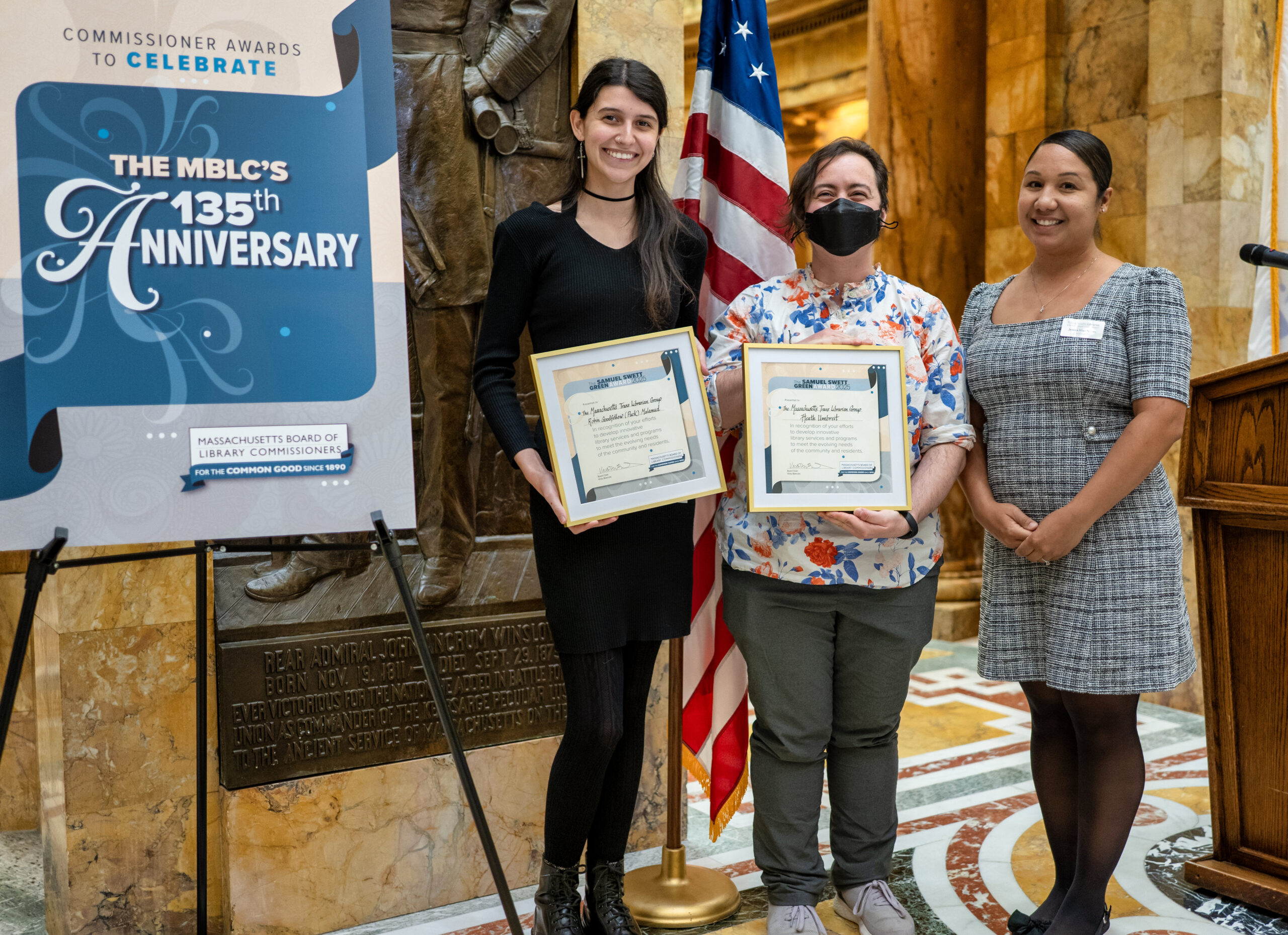 Three people stand indoors at the 2025 Commissioner Awards Ceremony, smiling. Two people are holding a framed certificate positioned next to a large sign reading “The MBLC’s 135th anniversary” with the “Massachusetts Board of Library Commissioners” logo below.