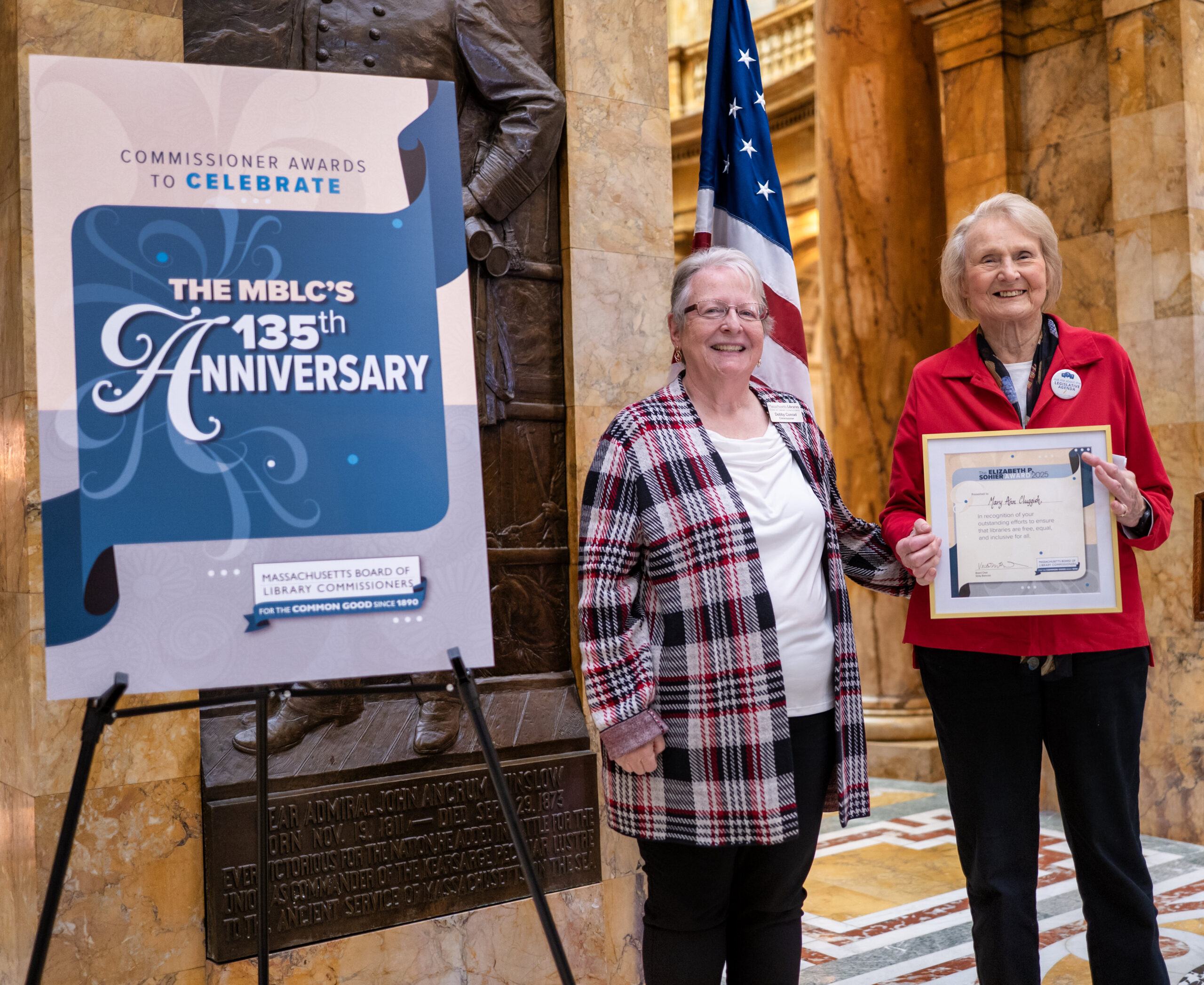 Two women stand indoors at the 2025 Commissioner Awards Ceremony, smiling and holding a framed certificate positioned next to a large sign reading “The MBLC’s 135th anniversary” with the “Massachusetts Board of Library Commissioners” logo below.